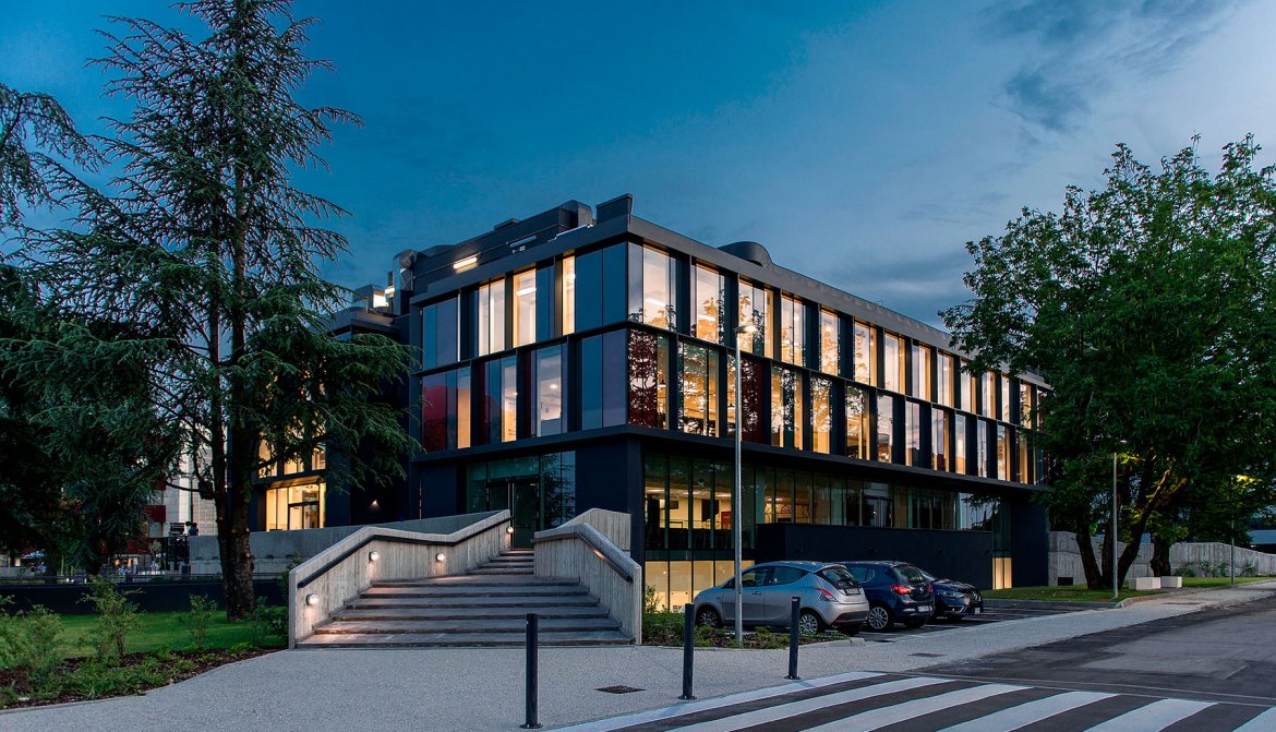 Edificio moderno per uffici con grandi finestre illuminate al crepuscolo, parcheggio con auto e alberi verdi intorno.