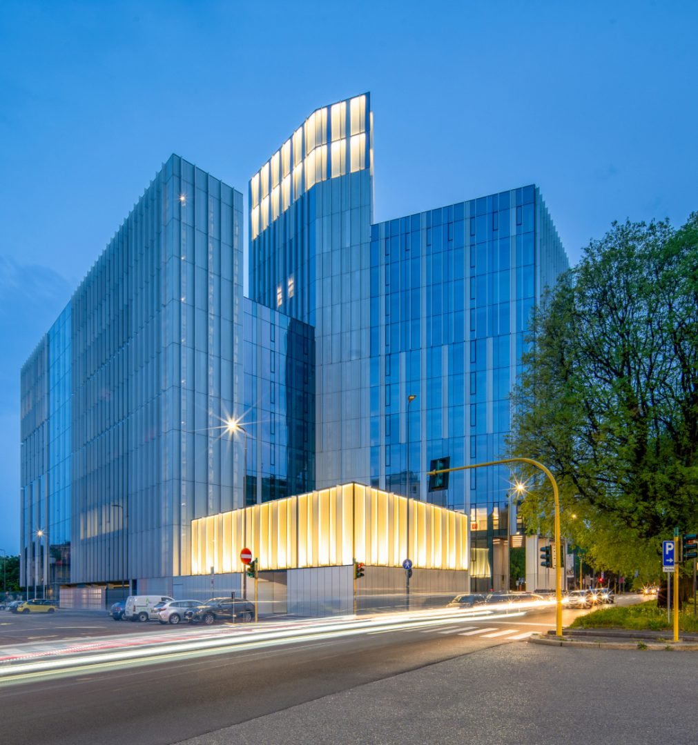 Edificio moderno con facciata in vetro illuminata di sera situato all'angolo di una strada trafficata con luci delle auto in movimento.