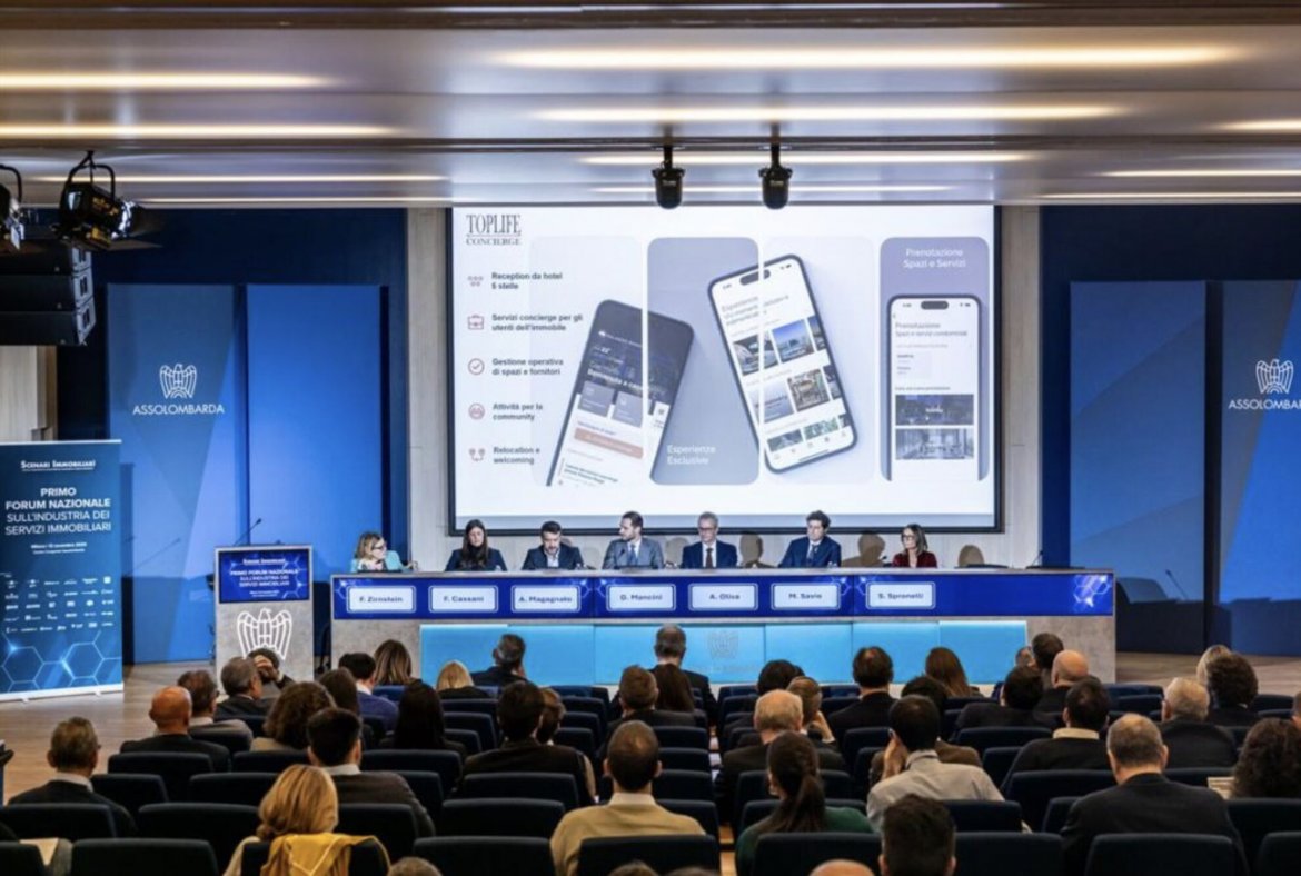 Conferenza con sette relatori seduti a un tavolo sul palco e pubblico in platea, con schermo che mostra un'applicazione mobile di servizi immobiliari.