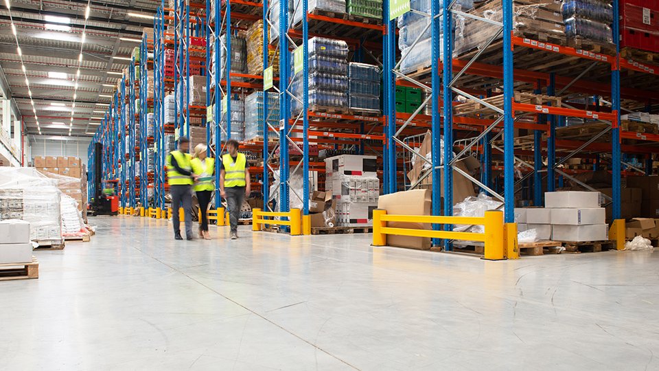 Tre lavoratori con giubbotti gialli camminano in un ampio magazzino con scaffalature alte piene di scatole e pallet.